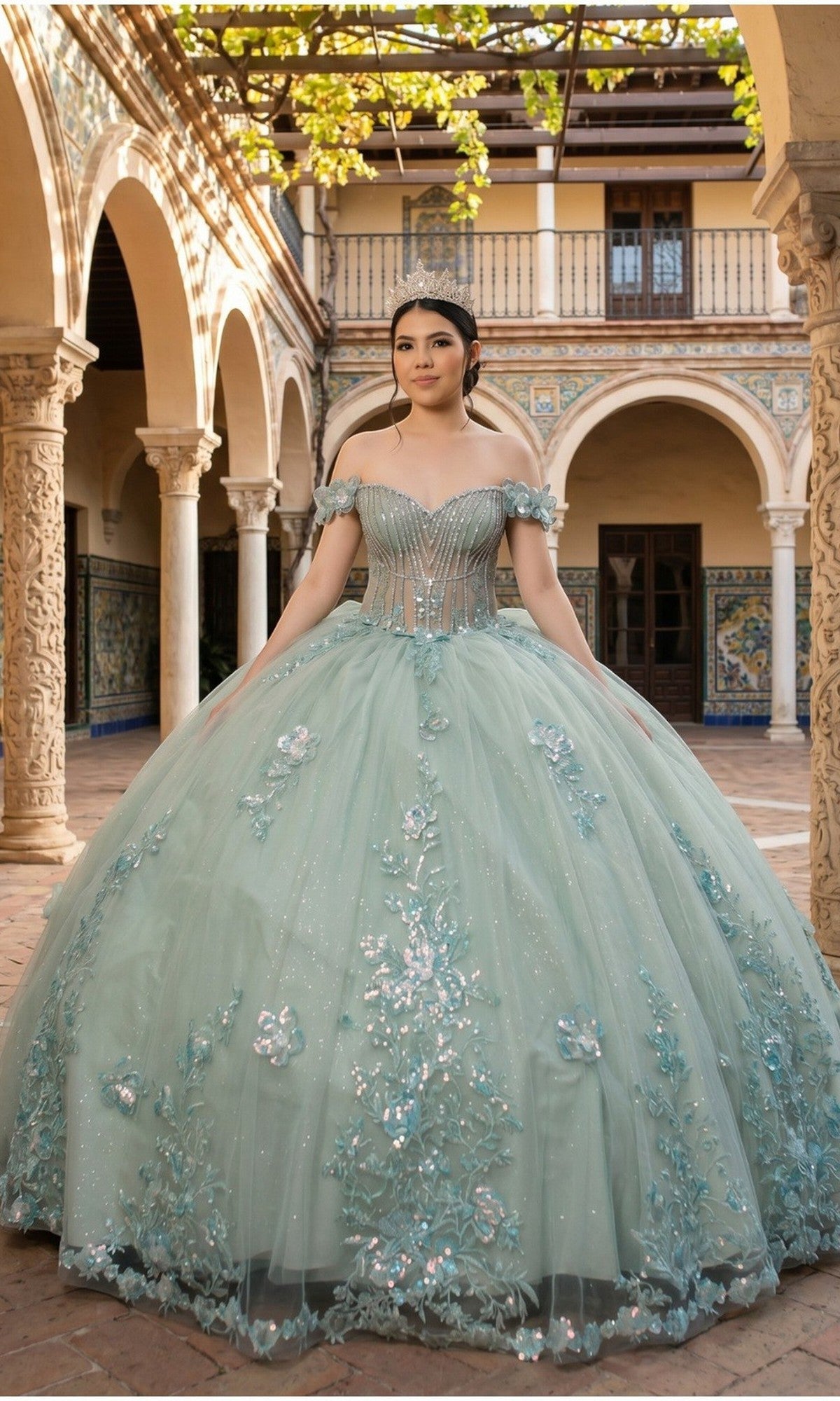 A young woman stands in a courtyard wearing the Quinceanera Dress 8370J by Cinderella Couture, featuring a sage hue, an off-the-shoulder neckline, delicate floral appliques, and a full ball gown skirt.